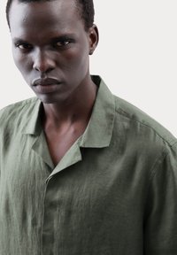 Groene linnen blouse met een ontspannen kraag, gestructureerde stof en een licht kreukelig uiterlijk. Close-up van de blouse gedragen door een model.