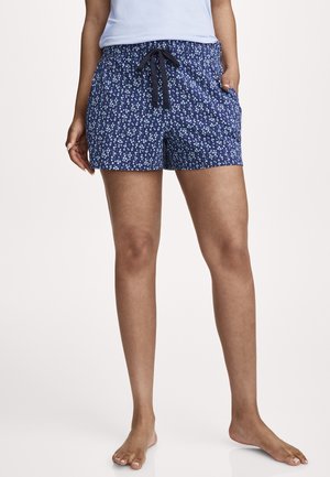 Woman wearing blue floral print shorts with black drawstring, paired with a light blue top, standing barefoot on a light background.