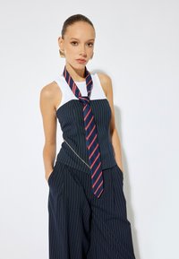 Blusa a rayas azul marino con cuello blanco, que presenta una bufanda de rayas atada en rojo y azul, combinada con pantalones holgados a juego en azul marino. Pendientes de aro dorados.