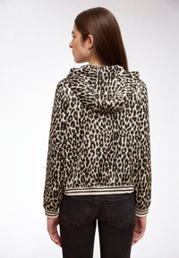 Frau, die rückwärts steht, trägt eine Kapuzenjacke mit Leopardenmuster sowie gestreiften Bündchen an Ärmeln und Saum, kombiniert mit schwarzer Jeans vor weißem Hintergrund.