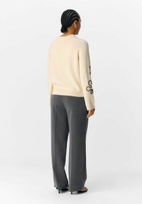 Maglione crema con ricami neri sulle maniche, abbinato a pantaloni grigi a gamba larga. Il materiale appare morbido con una finitura testurizzata.