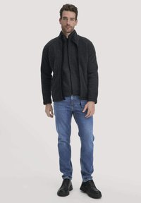 Dunkelgraue Zip-Up-Jacke mit hohem Kragen, getragen über einem schwarzen Shirt, kombiniert mit blauen Jeans und schwarzen Schuhen. Glatte Textur, lässige Passform.