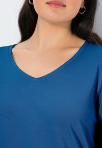 Niebieski t-shirt z dekoltem w serek, wykonany z miękkiej bawełnianej mieszanki tkanin. Charakteryzuje się gładką fakturą oraz widocznym przeszyciem wzdłuż linii dekoltu.