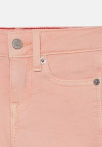 Różowy materiał dżinsowy z przednią kieszenią, metalowym zapięciem na guzik i widocznym przeszyciem. Gładka tekstura i luźny krój.