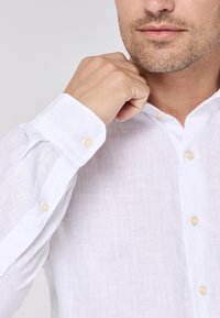 Weißes Leinenhemd mit Button-Down-Kragen, Einzelknopfmanschetten und subtiler strukturiertem Gewebe. Akzentuiert mit hellbraunen Knöpfen.