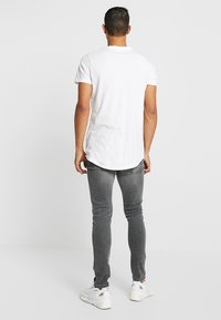 Vit kortärmad t-shirt med böjd nederkant, bärs med grå slim-fit jeans och vita sneakers, sedd från baksidan.