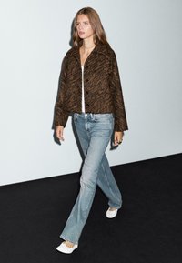 Veste cropped marron à motifs avec grand col, fermeture à boutons ; associée à un jean flare bleu clair et des ballerines blanches. Tissu lisse et texturé.