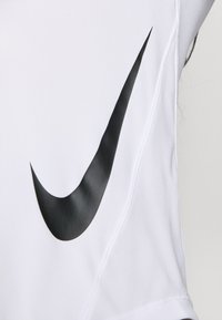 Biely atletický top s veľkým čiernym logom Nike Swoosh na ľavej strane. Materiál sa zdá byť hladký s priedušnou textúrou.