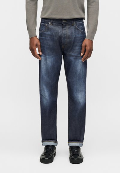 Άνδρας που φοράει σκούρα μπλε τζιν με γυρισμένα μπατζάκια, μαύρα αθλητικά παπούτσια και γκρι μπλούζα με μακριά μανίκια, στέκεται μπροστά σε ένα απλό φόντο.