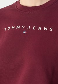 Sweatshirt bordeaux avec texte "TOMMY JEANS" brodé en blanc et petit logo Tommy Hilfiger centré en dessous du texte.