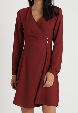 Nainen, jolla on pitkähihainen, polveen ulottuva viininpunainen kietaisumekko, jossa on V-aukko ja koristeelliset helmipainikkeet sivulla.