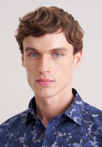 Niebieska koszula w kwiaty z kołnierzykiem na guziki, zdobiona ciemniejszymi kwiatowymi wzorami, wykonana z gładkiej, lekkiej tkaniny.