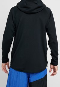 Chaqueta negra con capucha y superficie texturizada, con manga larga y aberturas laterales. La prenda se combina con pantalones cortos azules y negros.