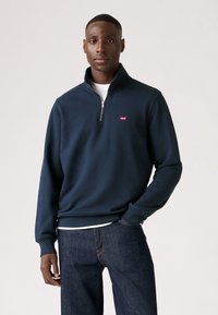 Marineblauwe zip-up sweatshirt van zachte stof, met een hoge kraag en een klein rood logo op de linkerborst. Gedragen met donkere jeans.