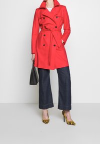 Trench-coat rouge à double boutonnage avec ceinture, associé à un jean large foncé et des talons imprimés tigre jaunes. Sac à main noir à la main.