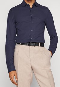 Camisa de manga larga azul marino con cierre de botones y cuello puntiagudo, combinada con pantalones beige claros y un cinturón negro con hebilla metálica.