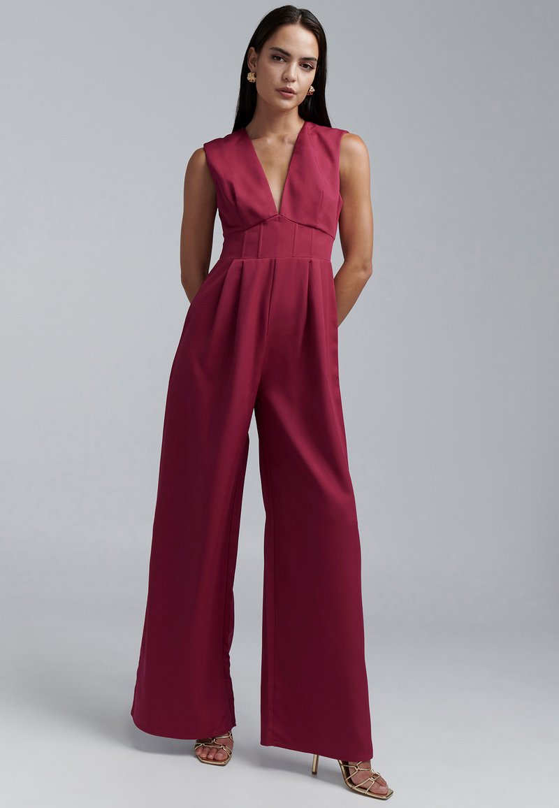 Mono sin mangas marrón con escote en V, diseño plisado y piernas anchas, confeccionado en tela suave, combinado con tacones dorados con tiras.