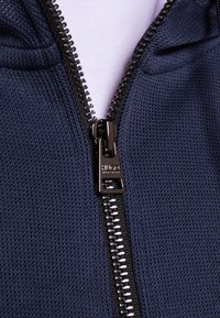 Marineblaue Zip-Hoodie aus strukturiertem Strickstoff, mit einem schwarzen Reißverschluss mit silbernen Zähnen und einem glatten Zipper.