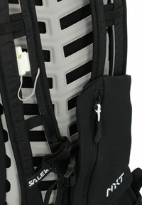 Spallacci di uno zaino da trekking nero con pannello posteriore in rete imbottita, piccola tasca con zip etichettata "MXT" e fischietto di sicurezza bianco attaccato.