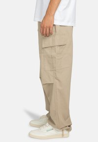 Pantaloni cargo beige in cotone con una vestibilità rilassata, caratterizzati da diverse tasche e dettagli cuciti, abbinati a sneakers bianche.