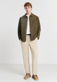 Camicia verde oliva con bottoni, dotata di tasca sul petto, abbinata a pantaloni beige e scarpe slip-on in suede color beige, su uno sfondo neutro.