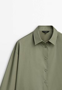 Chemise vert olive à manches longues, boutonnée, avec col pointu, présentée sur fond blanc.