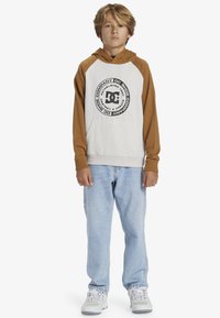 Sudadera con capucha de cuerpo gris y mangas marrones, con un logotipo negro circular. Combinada con unos jeans azul claro y zapatillas blancas.