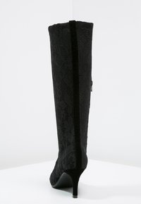 Stivale nero al ginocchio con texture in pizzo, tacco a stiletto e un pannello lucido lungo il retro. Presenta una chiusura con zip laterale.
