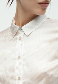 Πουκάμισο με γιακά σε ανοιχτό ροζ σατέν με κάθετες ρίγες· διαθέτει διακοσμητικό κουμπί με ριντόν και πέρλες κατά μήκος του μπροστινού μέρους.