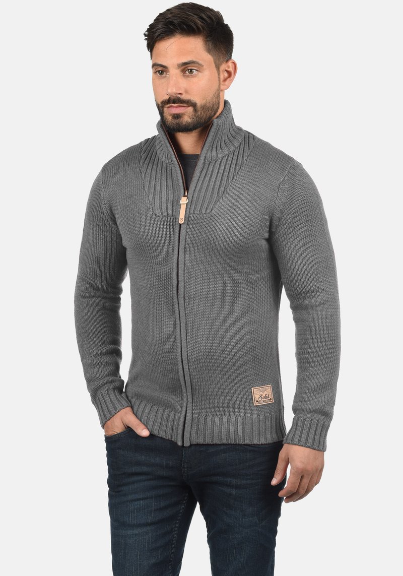 Graue Strickjacke mit hohem Kragen und Reißverschluss. Verfügt über gerippte Bündchen und Saum sowie ein kleines Leder-Logo-Patch unten links.