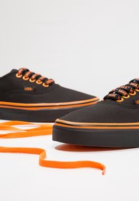 Des baskets noires en toile avec des lacets et des accents orange, dotées d'une semelle en caoutchouc et d'une bande orange. Les lacets sont disposés à côté des chaussures.