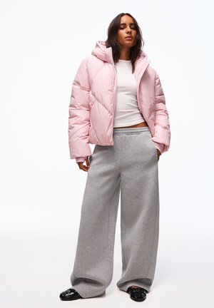 Femme portant une doudoune rose clair, un t-shirt blanc court, un pantalon de survêtement large gris et des chaussures noires, debout les mains dans les poches.