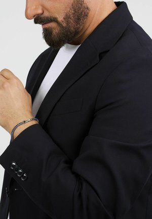 Blazer nero con spalle strutturate e revers, con una singola tasca sul petto. Il polsino presenta tre bottoni e un braccialetto intrecciato al polso.