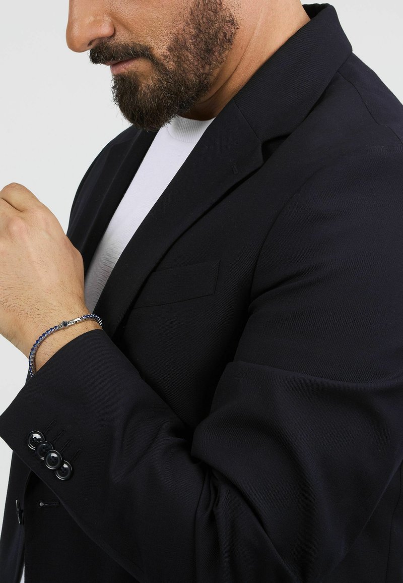 Blazer nero con spalle strutturate e revers, con una singola tasca sul petto. Il polsino presenta tre bottoni e un braccialetto intrecciato al polso.