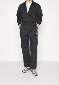 Svart skjorta med knappar och två bröstfickor, matchad med svarta byxor med elastisk midja som har en Nike-logotyp, och grå sneakers.