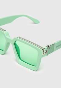 Gafas de sol cuadradas verde menta con lentes tintados, montura de plástico grueso y detalles metálicos. Las patillas cuentan con un detalle en forma de corazón.