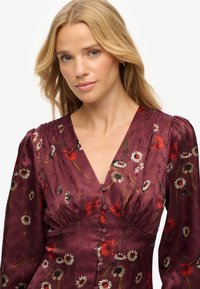Vestido floral en burdeos profundo con escote en V, mangas largas abullonadas y detalles de botones. Presenta amapolas rojas y margaritas blancas sobre una textura sedosa.