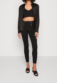 Camisa de satén negra superpuesta sobre un top corto negro, combinada con leggings de cintura alta con detalles de botones metálicos y tacones negros deslizables.