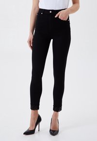 Jeans negros de talle alto con un diseño ajustado, que presentan detalles de encaje en el tobillo y dos bolsillos frontales. Llevados con tacones altos negros.