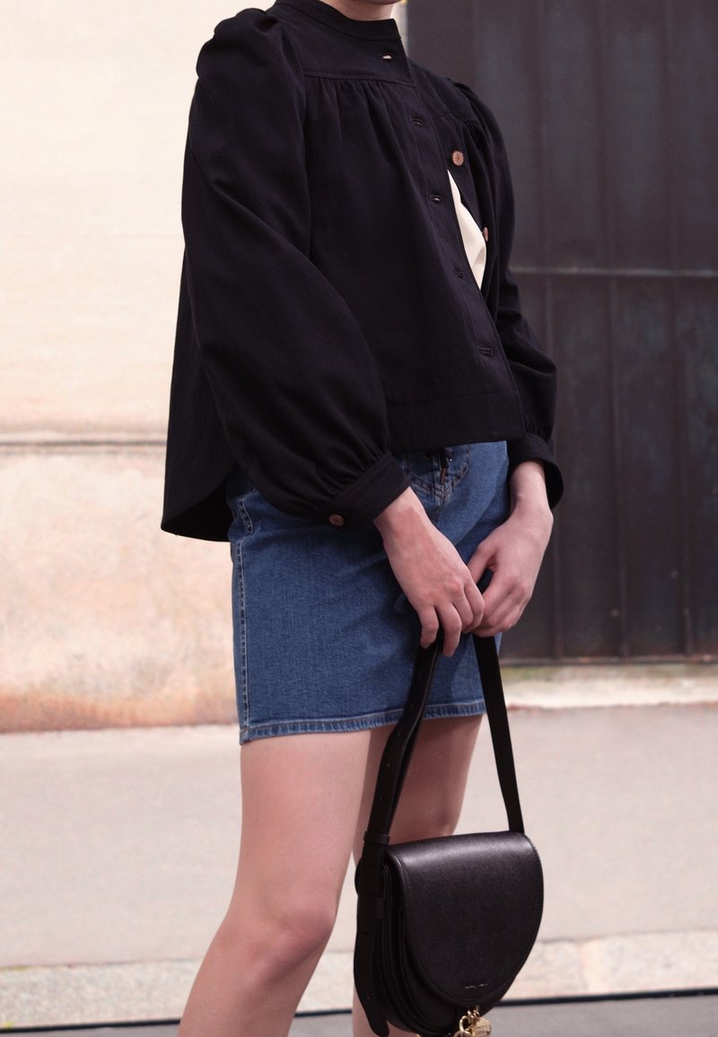 Personne portant un chemisier noir à manches longues et une jupe en jean bleue, tenant un petit sac à main noir avec les deux mains.