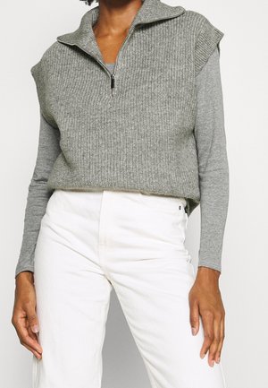 Persoon die een grijs, mouwloos gebreid vest met een rits aan de voorkant draagt over een shirt met lange mouwen en een witte broek met hoge taille.