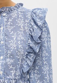 Blouse en vichy bleu et blanc avec broderie florale blanche et détail de manches à volants, avec des boutons blancs sur le devant.