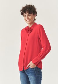 Blusa roja de manga larga con un detalle de rosa en el cuello, hecha de una tela suave. Combinada con jeans de mezclilla azules.