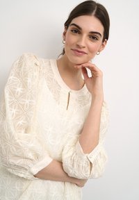 Krémová blůza s výšivkou a nabíranými rukávy. Disponuje výstřihem do tvaru klíče a knoflíkovými detaily. Látka je průsvitná s květinovými vzory.