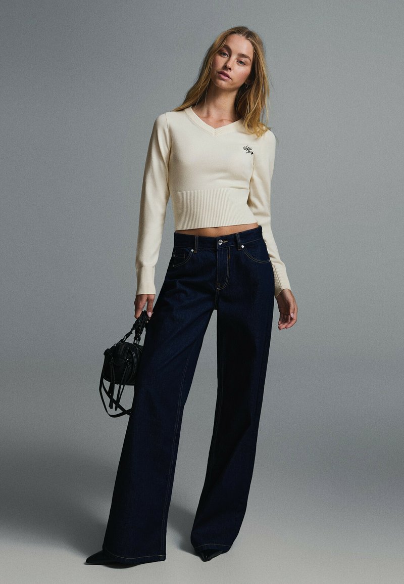 Femme portant un pull court crème à manches longues et un jean large foncé, tenant un petit sac à main noir sur un fond gris uni.