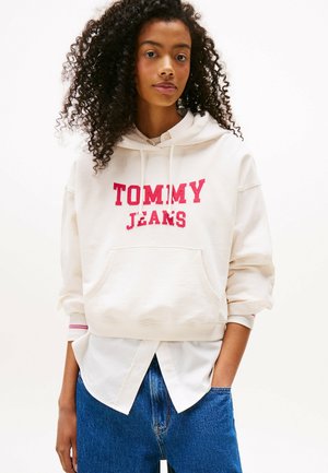 Crème hoodie met "TOMMY JEANS" in rode letters, met een voorzak en opgerolde mouwen over een wit shirt en blauwe jeans.