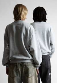 Felpa grigia con maniche lunghe e polsini a costine, abbinata a jeans baggy scuri. Vista frontale e posteriore di due individui in piedi ravvicinati.