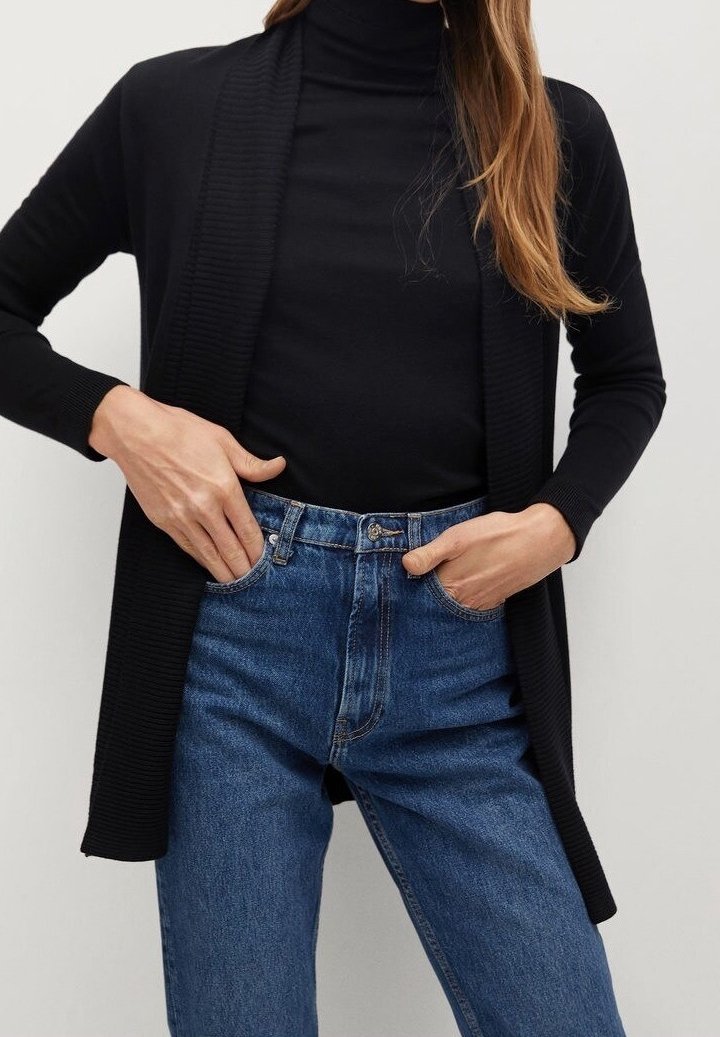 Femme portant un col roulé noir à manches longues et un cardigan noir ouvert, associé à un jean bleu taille haute, mains dans les poches.