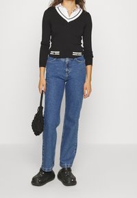 Pull en maille noir avec un col en V blanc et des accents rayés, associé à un jean droit bleu et des sabots noirs, tenant un sac noir.