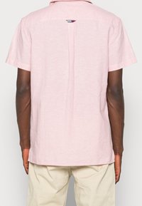 Uomo che indossa una camicia leggera rosa a maniche corte e pantaloni beige, visto da dietro su uno sfondo neutro.